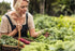 A blonde woman in overalls harvesting fresh green produce.